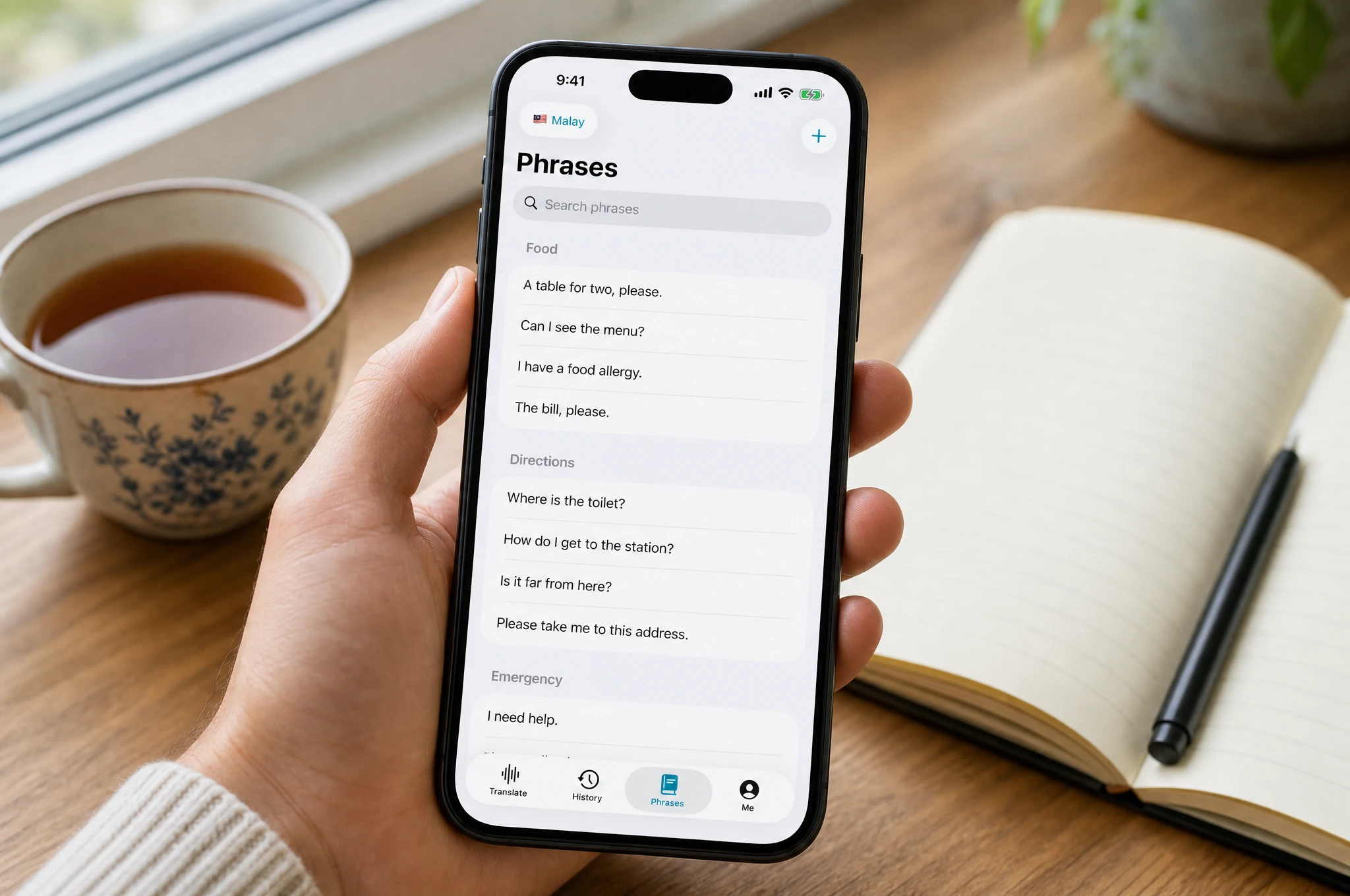 Hand holding a smartphone showing the Nusan phrasebook set to Malay, with categorized everyday phrases