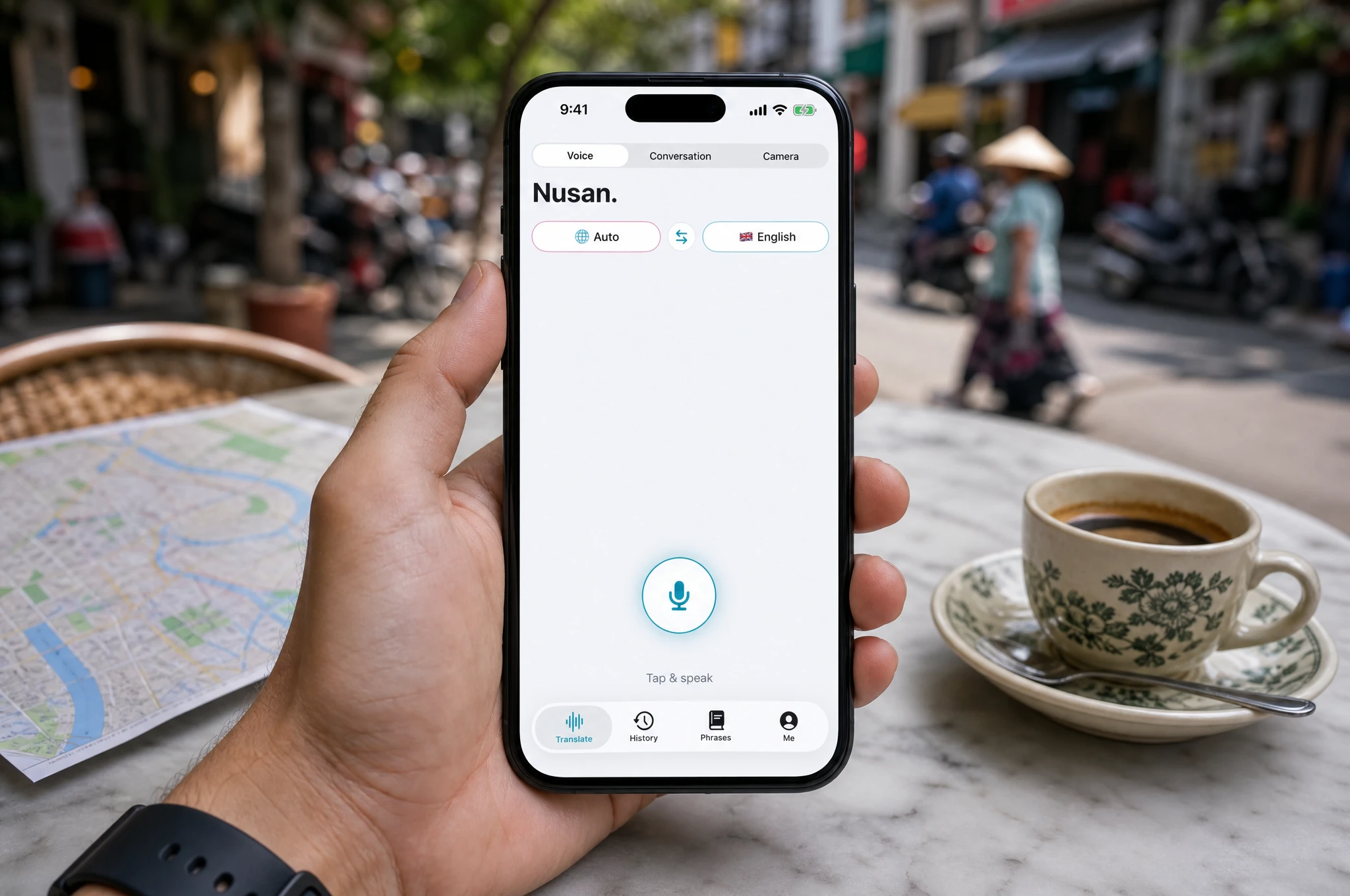 Hand holding a smartphone at an outdoor cafe table in Vietnam, showing the Nusan voice translation screen ready to listen