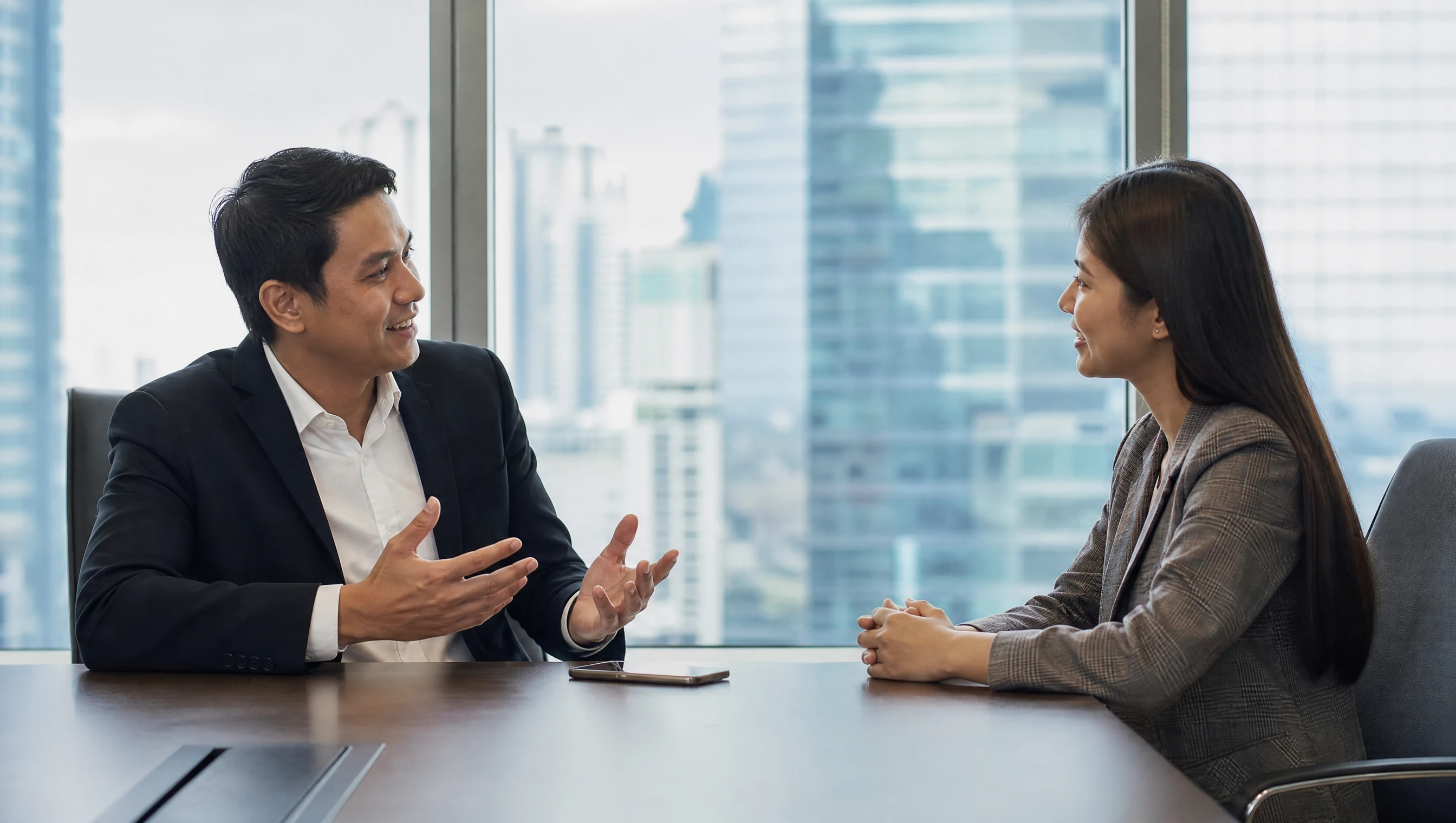 Two professionals in friendly conversation across a meeting table, a smartphone between them