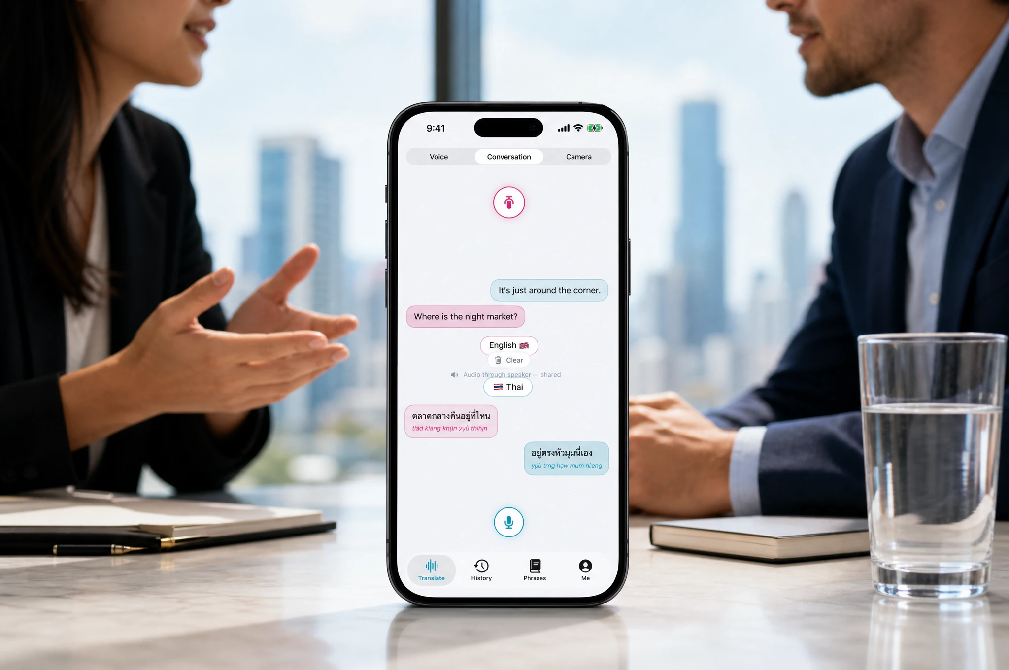 Smartphone standing on a meeting table showing the Nusan conversation mode with a Thai and English exchange, business meeting in the background